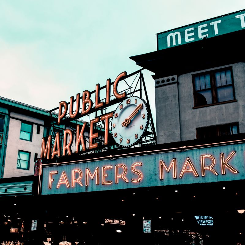 A historic public market with neon signs reading "Public Market" and "Farmers Market," featuring a clock and surrounded by old buildings.