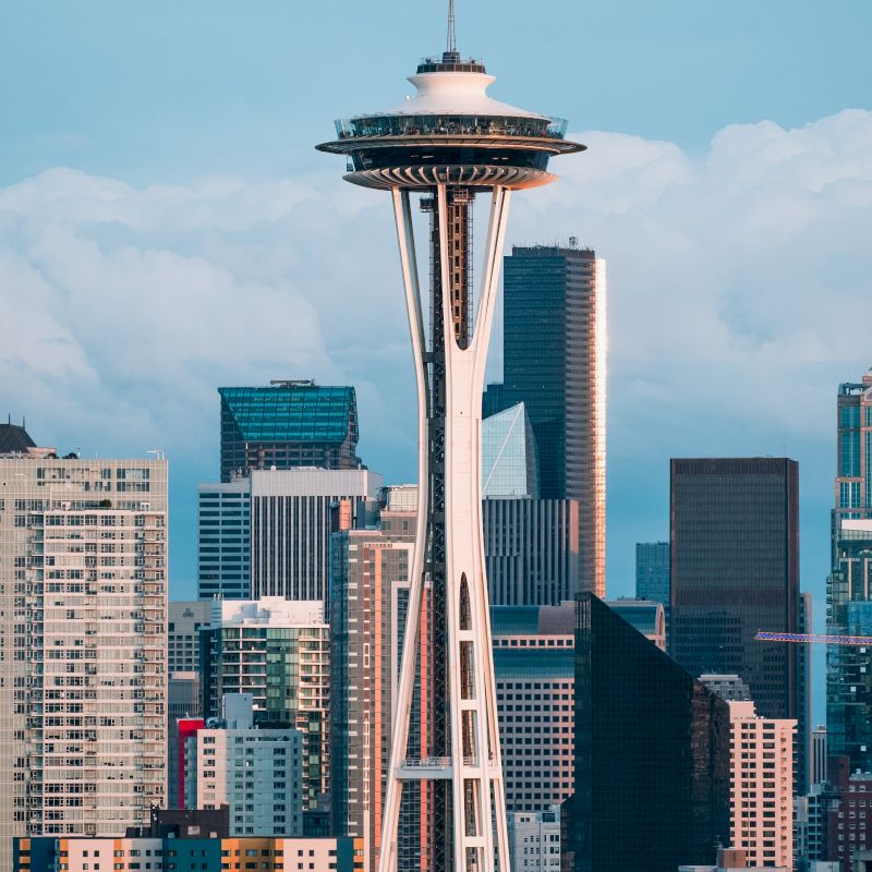 The image shows the Space Needle against a backdrop of various skyscrapers and buildings in what appears to be Seattle, with cloudy skies overhead.