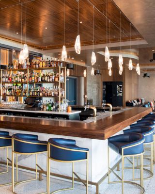 This image features a modern bar with blue stools, illuminated hanging lights, and a well-stocked bar counter, with a seating area in the background.