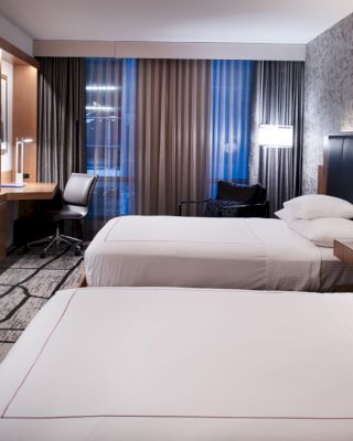 A modern hotel room with two beds, a flat-screen TV, a desk and chair by the window, nightstands with lamps, and patterned walls and carpet.