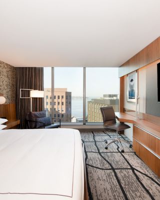 A modern hotel room with a large bed, wall-mounted TV, desk, and chair, featuring a city view through a large window.
