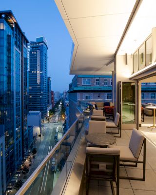 A modern cityscape at dusk viewed from a balcony with sleek furniture and buildings illuminated by city lights in the background.