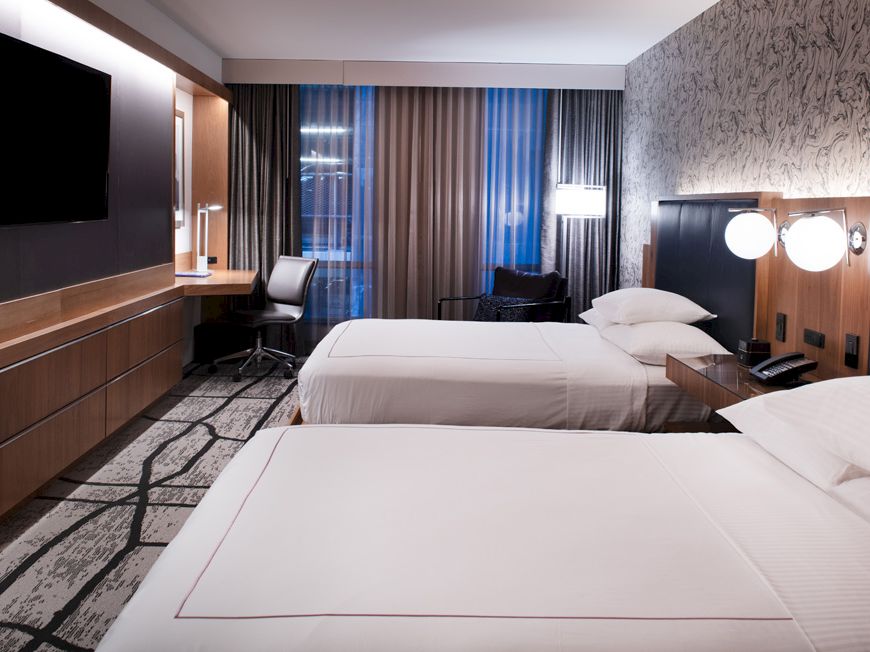 A modern hotel room with two neatly made beds, a flat-screen TV, a desk, and contemporary decor with patterned wallpaper and curtains.