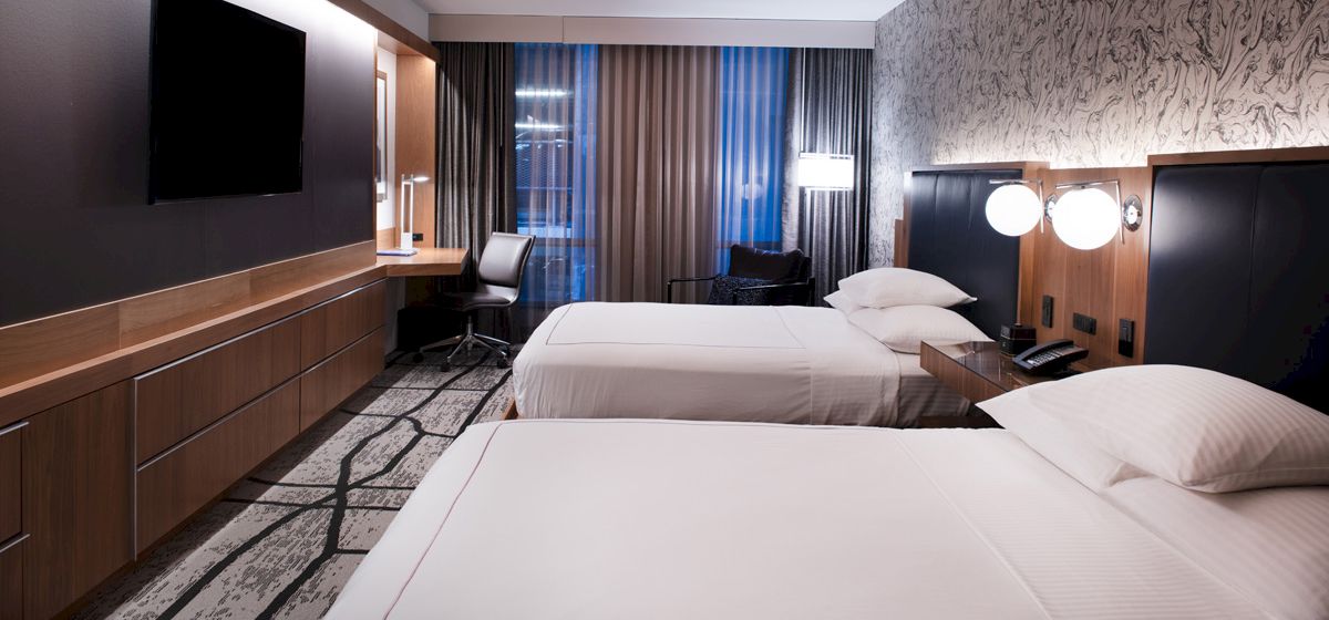 A modern hotel room with two neatly made beds, a flat-screen TV, a desk, and contemporary decor with patterned wallpaper and curtains.