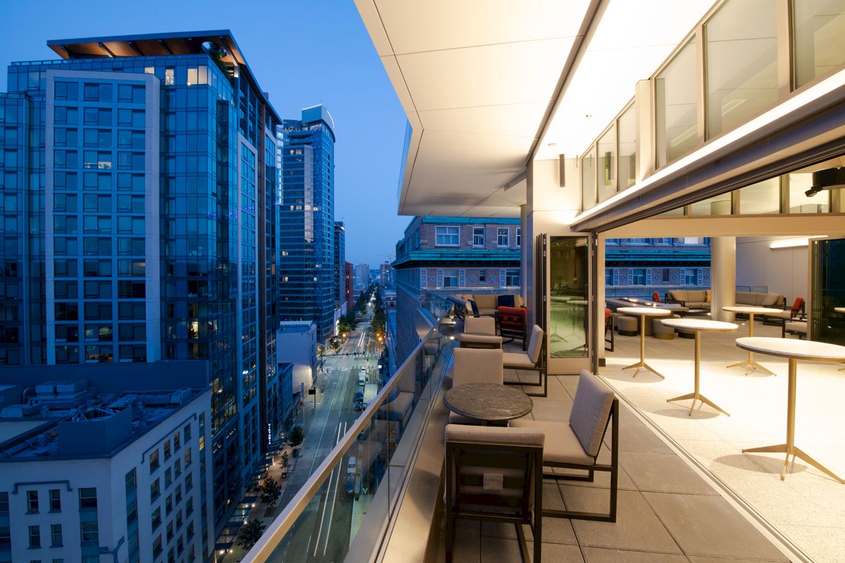 A modern urban balcony overlooks a cityscape at dusk. The balcony has outdoor seating with glass railings and merges into an indoor area.