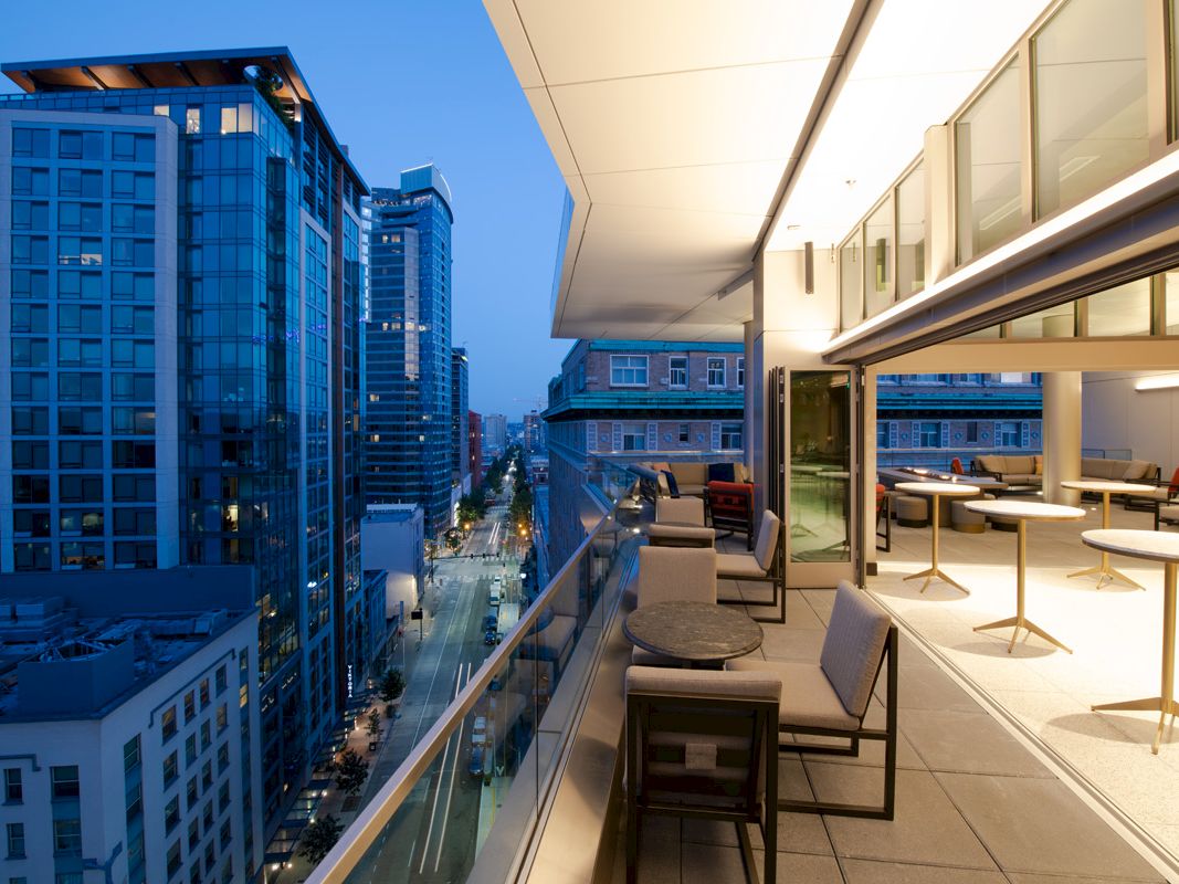 A modern urban balcony overlooks a cityscape at dusk. The balcony has outdoor seating with glass railings and merges into an indoor area.