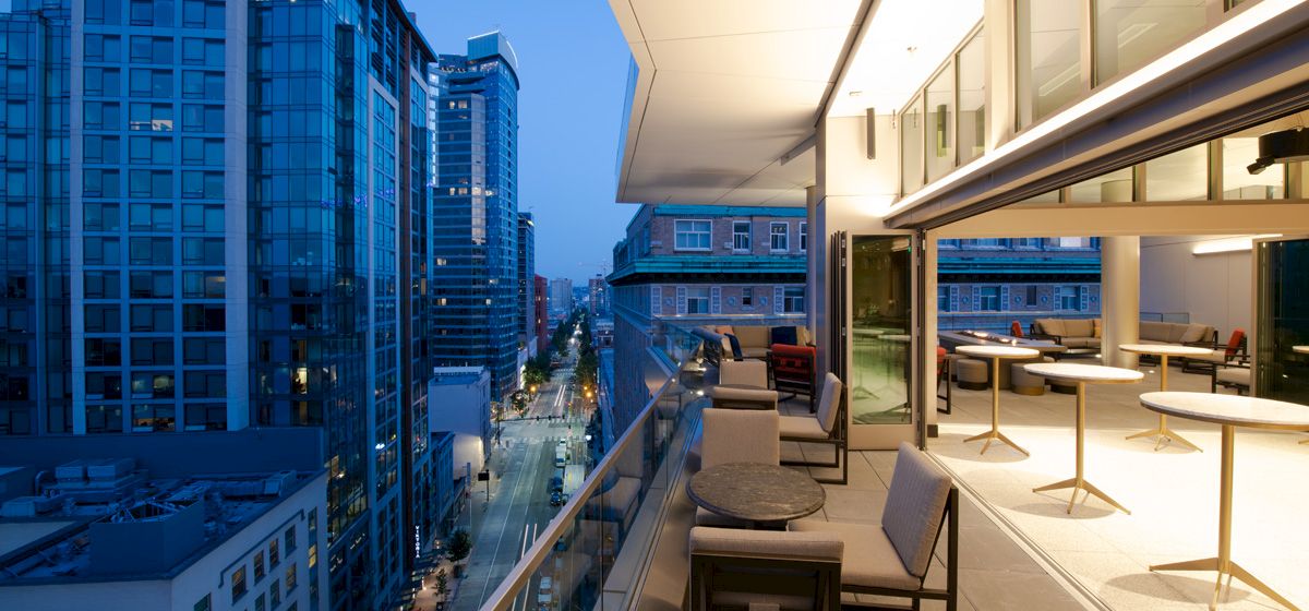 A modern urban balcony overlooks a cityscape at dusk. The balcony has outdoor seating with glass railings and merges into an indoor area.