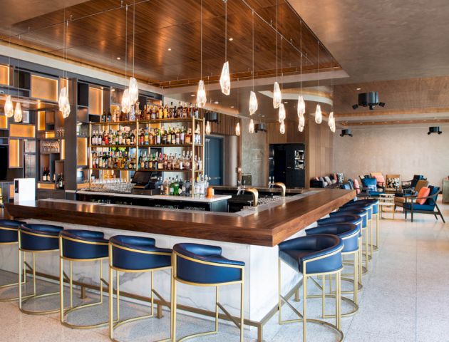 A stylish bar area with a wooden counter, blue chairs, hanging lights, and a fully stocked back bar; a seating area is visible in the background.