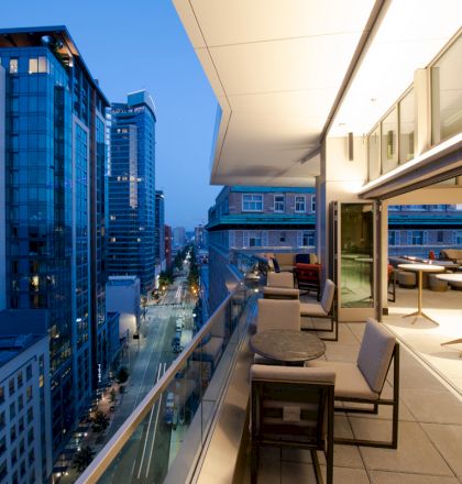 A modern, outdoor balcony view of a cityscape at dusk, adorned with tables, chairs, and building lights reflecting off nearby skyscrapers.