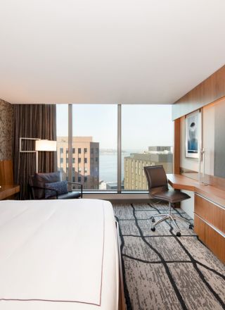 A modern hotel room with a large bed, wall-mounted TV, desk, chair, and a window offering a cityscape view with water in the background.