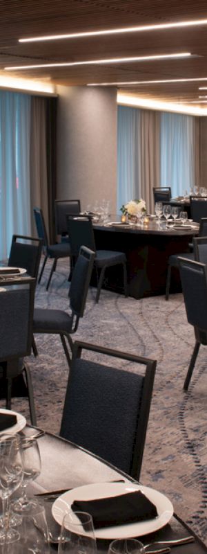 The image shows a well-set dining area with round tables and chairs, elegant table settings, and soft lighting. The space has floor-to-ceiling windows.