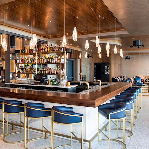A modern bar with blue and gold bar stools, a well-stocked counter, and contemporary lighting fixtures in a stylish, spacious setting.