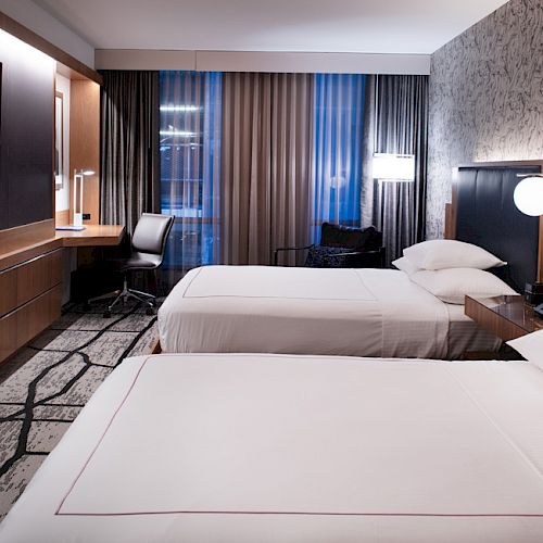 A modern hotel room with two beds, a flat-screen TV, desk, chair, and large windows with curtains. Walls feature a decorative pattern.