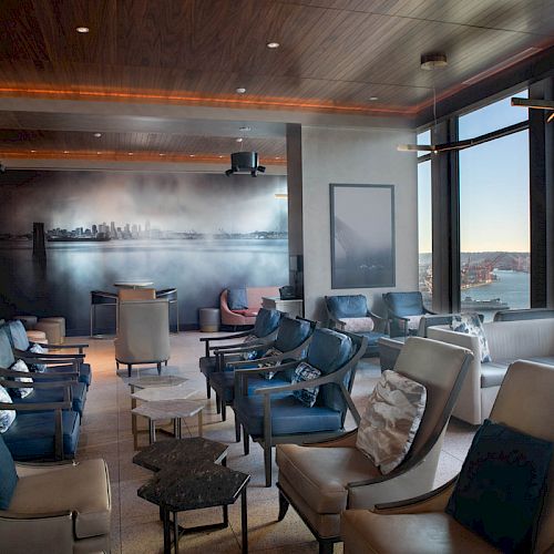 The image shows a modern, well-lit lounge area with comfortable seating, large windows with a scenic view, and a mural of a city skyline.