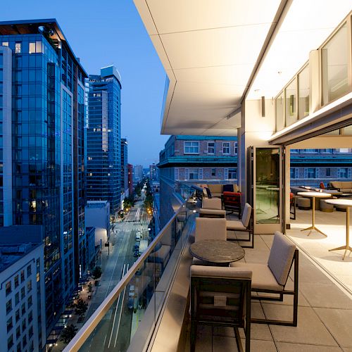 A modern cityscape at dusk featuring tall buildings and a stylish outdoor balcony with seating areas and tables overlooking the street.