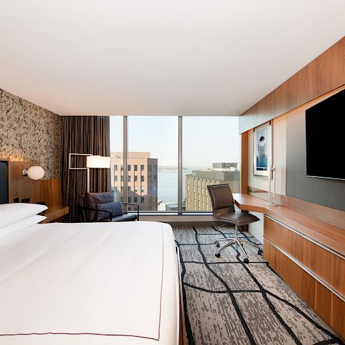 A modern hotel room features a large bed, wall-mounted TV, desk, and chair with windows revealing a cityscape view and abstract wall art.