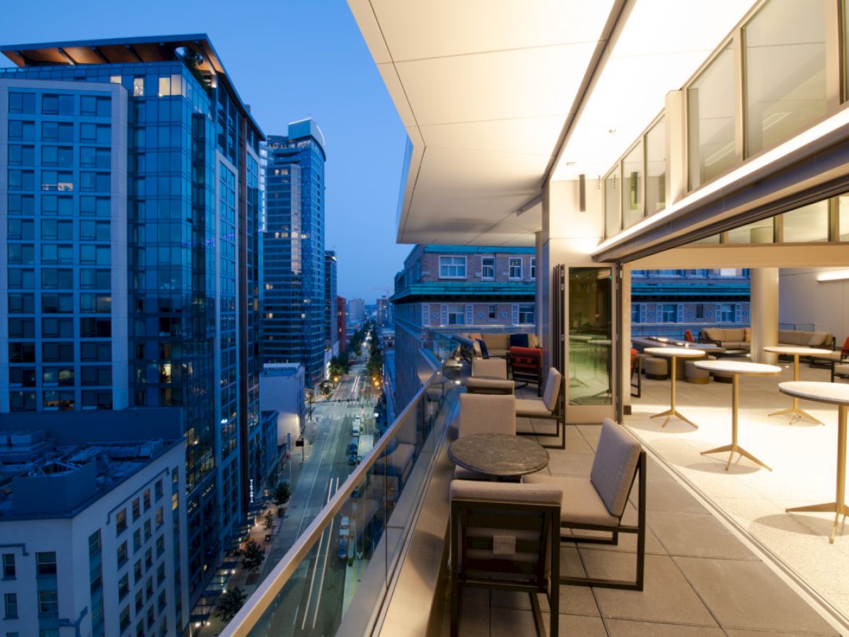 The image shows a modern balcony at dusk, overlooking a cityscape with tall buildings and streets below. The balcony has table seating and open glass doors.