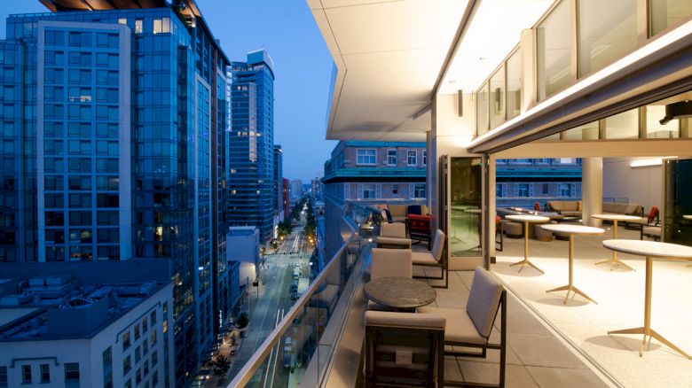 The image shows a modern balcony at dusk, overlooking a cityscape with tall buildings and streets below. The balcony has table seating and open glass doors.
