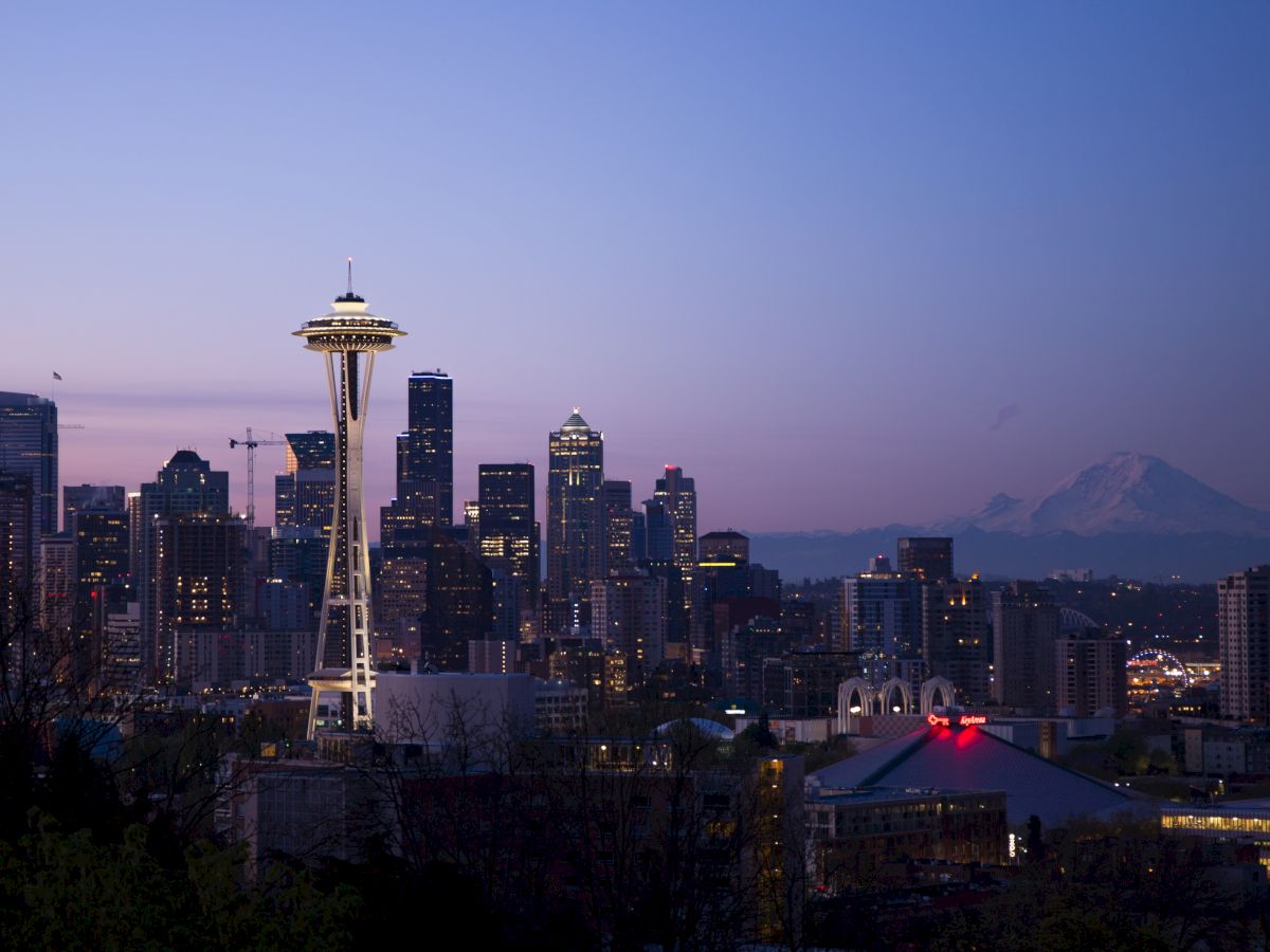The image shows the Seattle skyline at dusk, featuring the Space Needle prominently with Mount Rainier in the background and city lights beginning to illuminate.