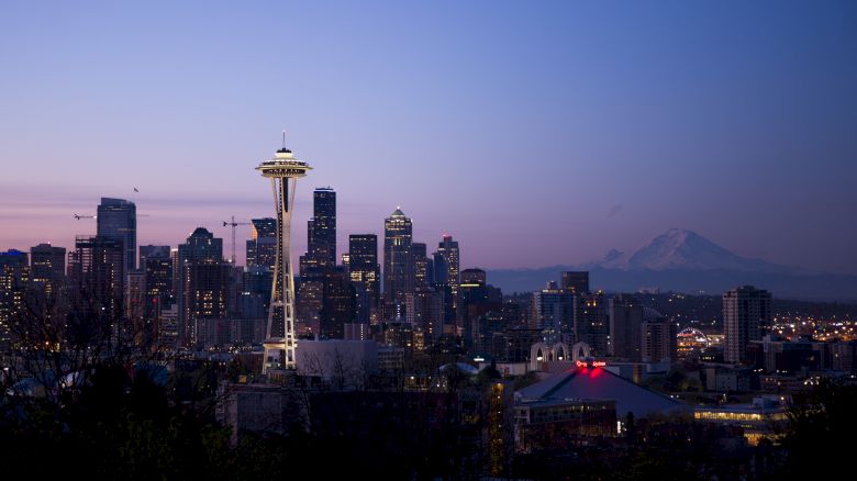 The image shows the Seattle skyline at dusk, featuring the Space Needle prominently with Mount Rainier in the background and city lights beginning to illuminate.