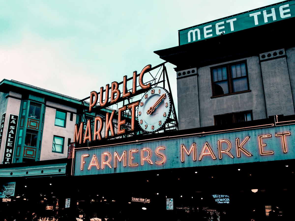 An iconic outdoor public market sign, featuring a clock, above a vibrant "Farmers Market" in neon, with adjacent buildings and a partially visible "MEET THE" sign.