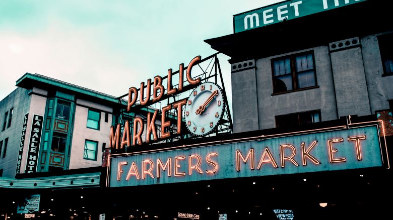 An iconic outdoor public market sign, featuring a clock, above a vibrant "Farmers Market" in neon, with adjacent buildings and a partially visible "MEET THE" sign.