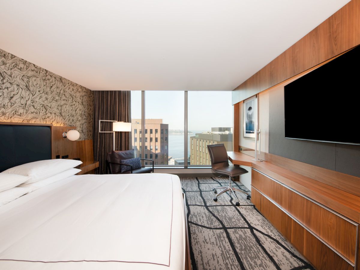 This is a modern hotel room featuring a large bed, desk with a chair, a wall-mounted TV, and a window with a view of buildings outside.