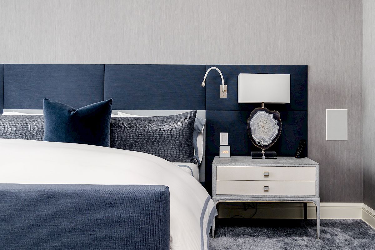 A modern bedroom with a blue and white color scheme, featuring a bed, blue headboard, bedside table, lamp, and decorative accessories.