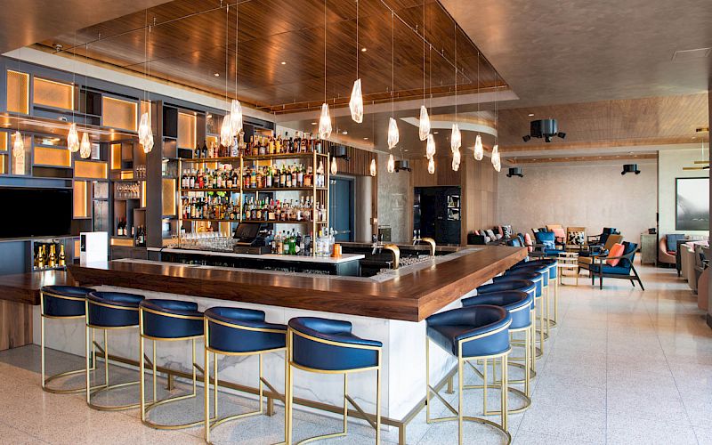 This image shows a stylish, modern bar with a wooden counter, blue chairs, hanging lights, and a well-stocked selection of drinks, set in a chic interior.