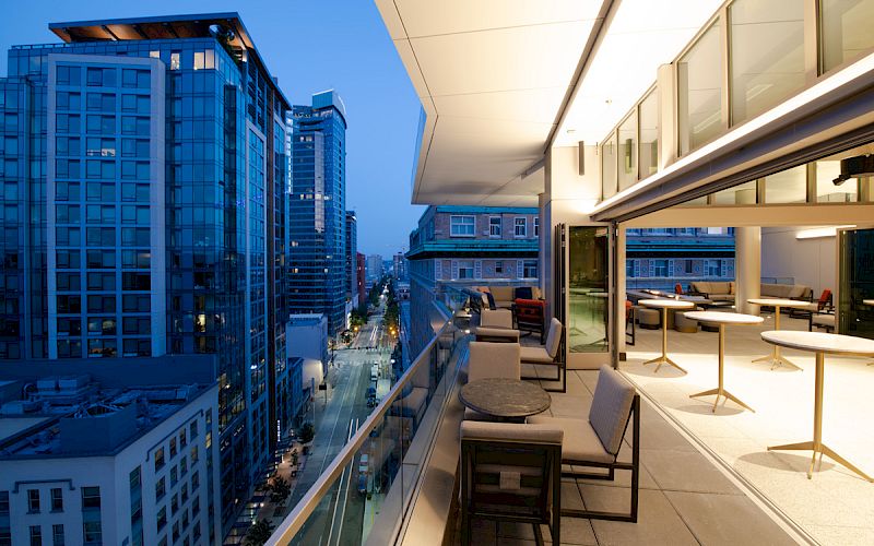 A modern cityscape at dusk with high-rise buildings and a residential balcony featuring seating and tables overlooking the urban scene.