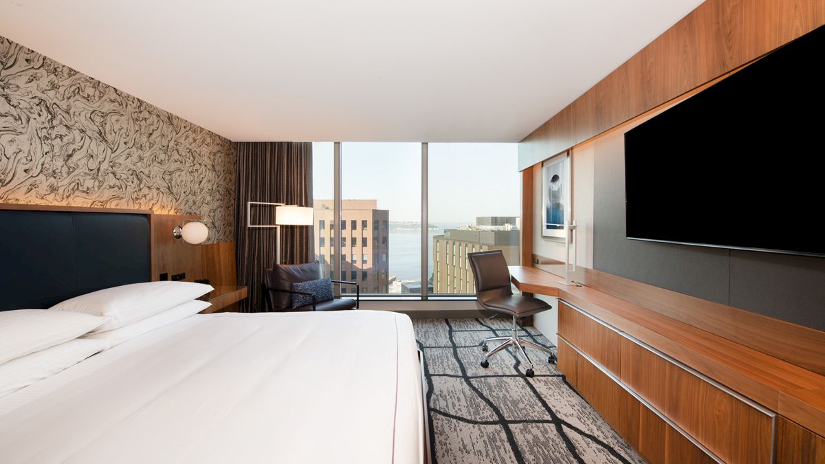 A modern hotel room with a large bed, desk, chair, and TV. There is a cityscape view through the large windows and contemporary decor.