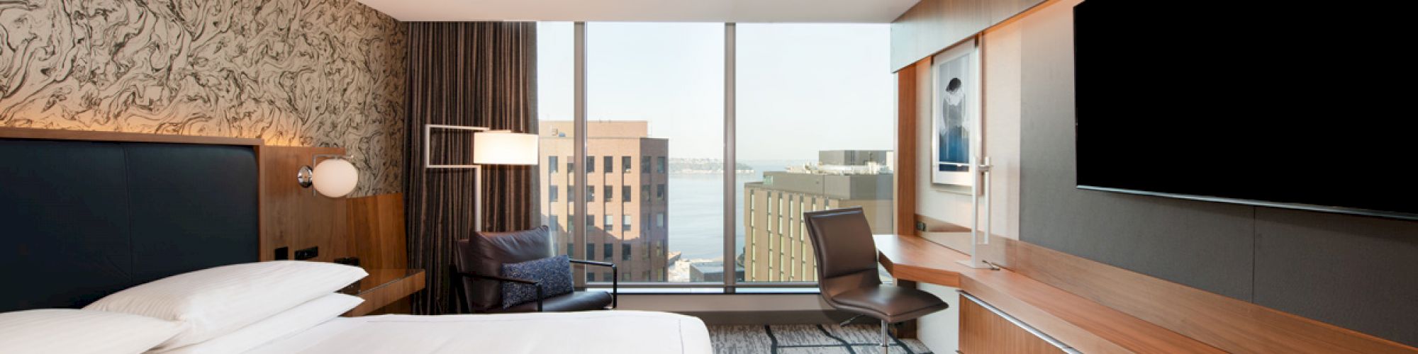 A modern hotel room with a large bed, desk, chair, and TV. There is a cityscape view through the large windows and contemporary decor.