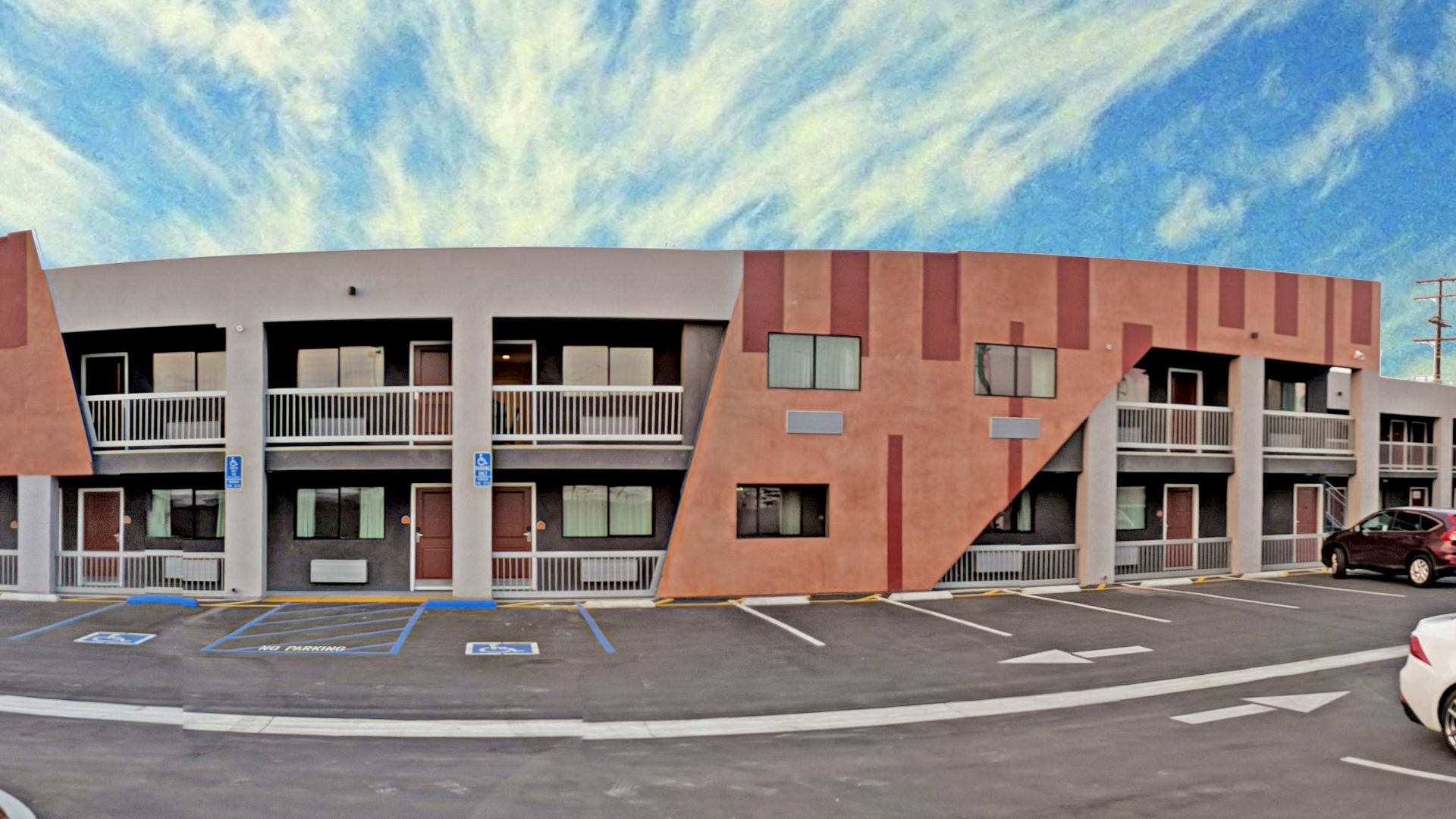 The image shows a two-story motel building with several parking spaces in front, some containing cars, under a sky with scattered clouds.
