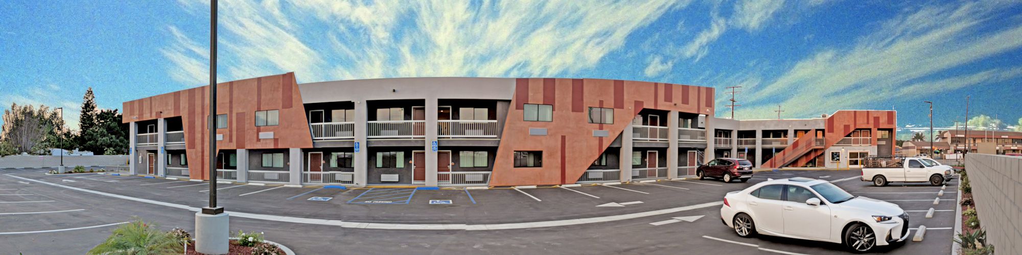 The image shows a two-story motel building with several parking spaces in front, some containing cars, under a sky with scattered clouds.