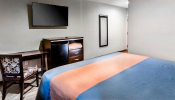 The image shows a hotel room with a bed, a desk, a TV mounted on the wall, a dresser, a refrigerator, and a mirror. The bed has blue and orange bedding.