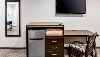 A hotel room setup with a mini-fridge, drawers, folded towels, a wall-mounted TV, desk, chair, and a full-length mirror.