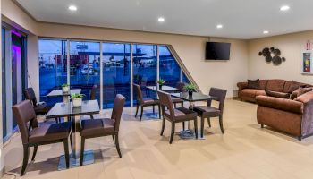 The image shows a modern lounge area with tables and chairs, a sofa set, a wall-mounted TV, and windows showcasing a view of outside buildings.