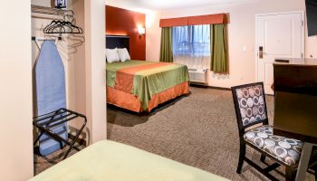 This image shows a hotel room with a bed, ironing board, chair, and desk. The room is well-lit with a window and doorway.