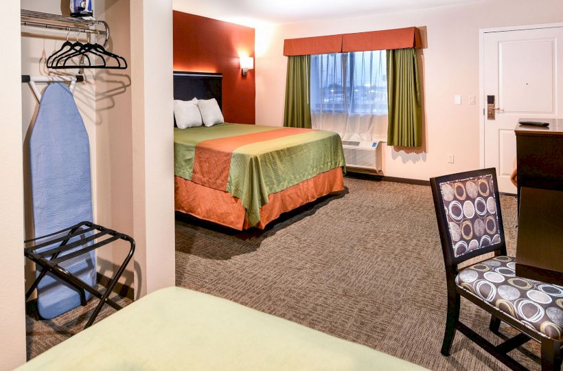 This image shows a hotel room with a bed, ironing board, chair, and desk. The room is well-lit with a window and doorway.