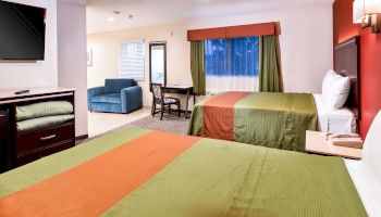 A hotel room with two beds, TV, sofa, table, and chairs, featuring green and orange bedding, and a window with curtains ending the sentence.