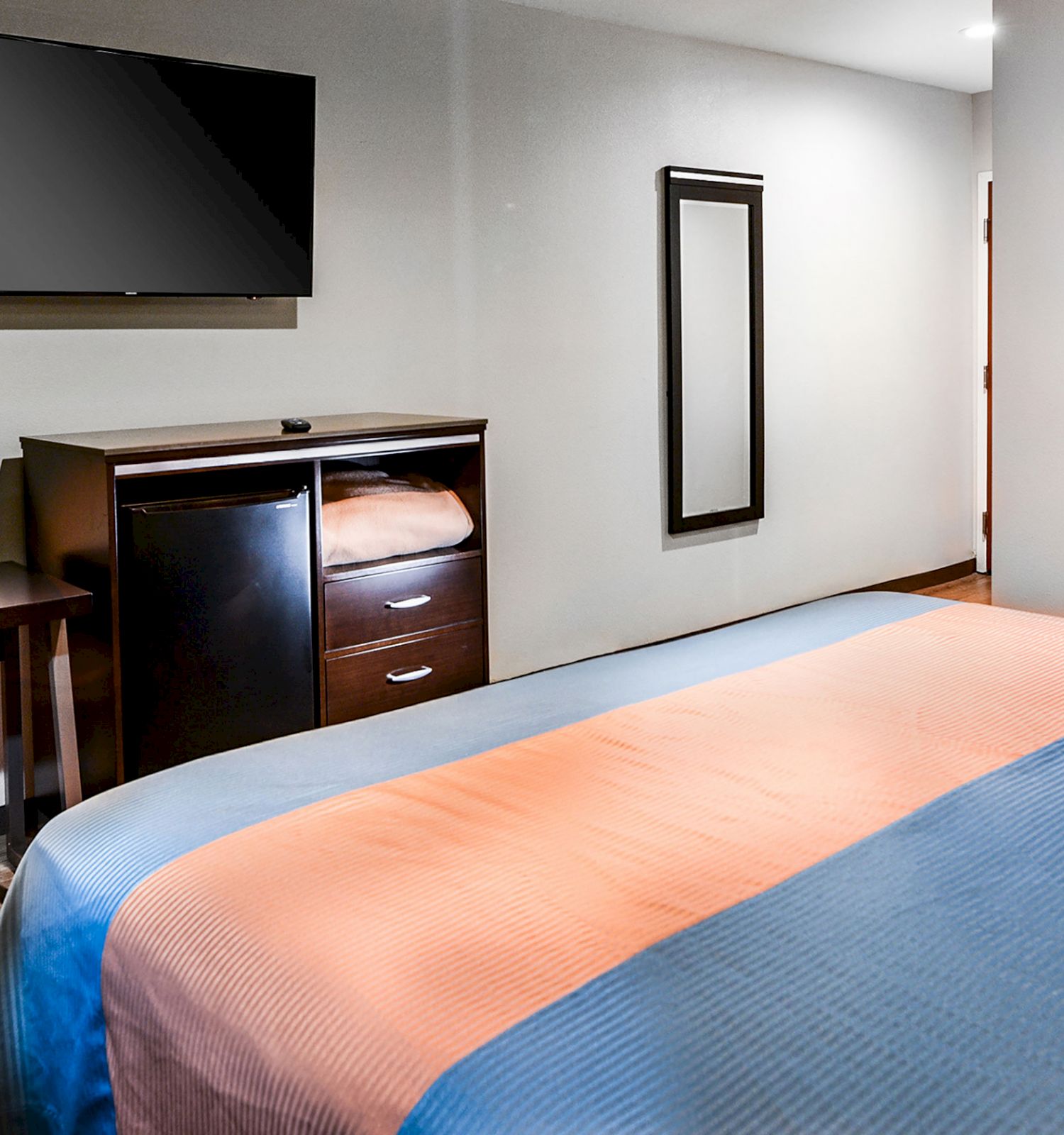 The image shows a modern hotel room with a neatly made bed, a wall-mounted TV, a desk with a chair, and a dresser beneath the TV.