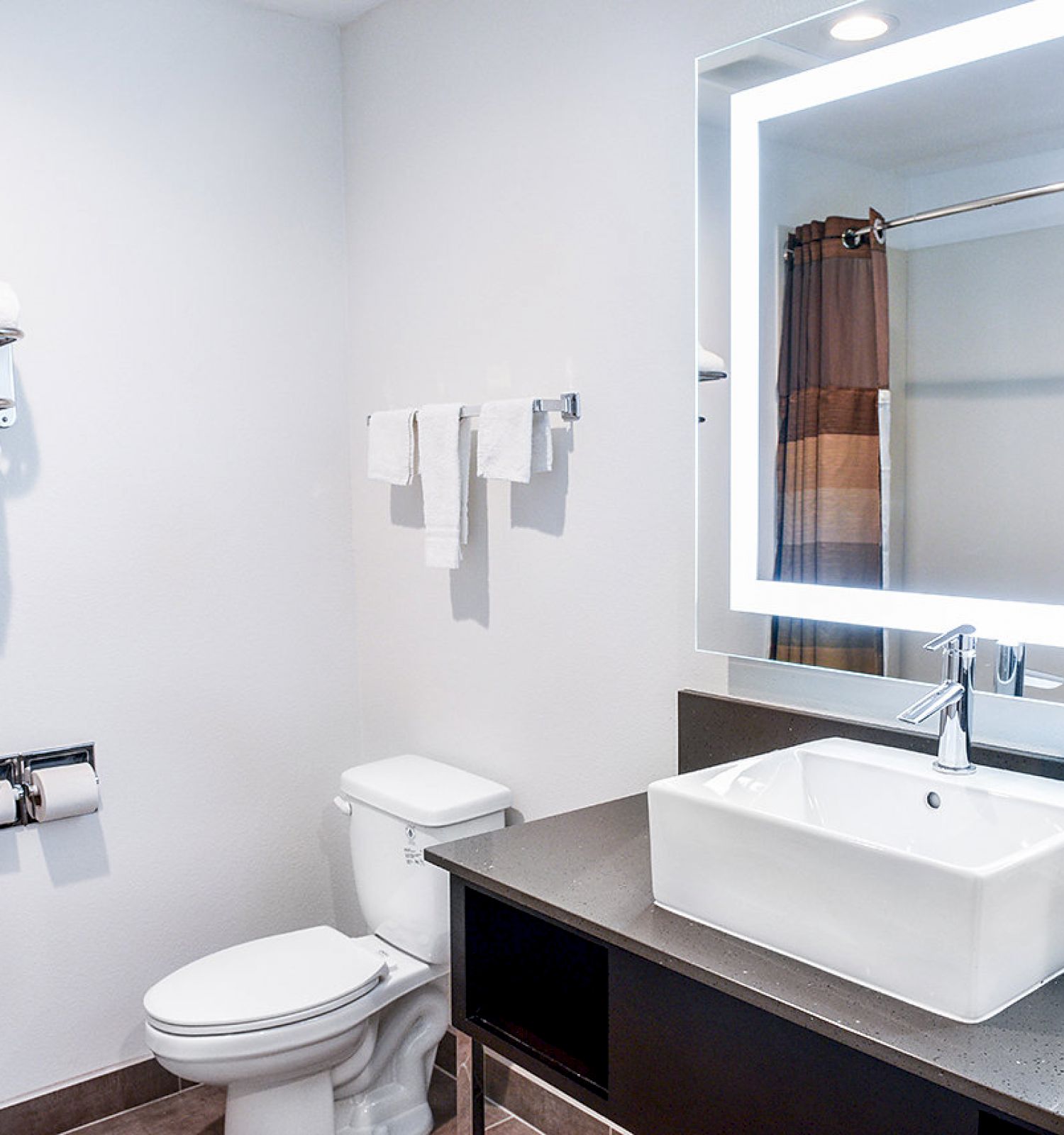 A modern bathroom with a lighted mirror, white sink, toilet, towels on racks, and a shower with a curtain.