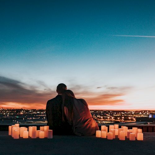 A couple sitting close together on a rooftop at sunset, surrounded by lit candles, overlooking a cityscape with a colorful sky.