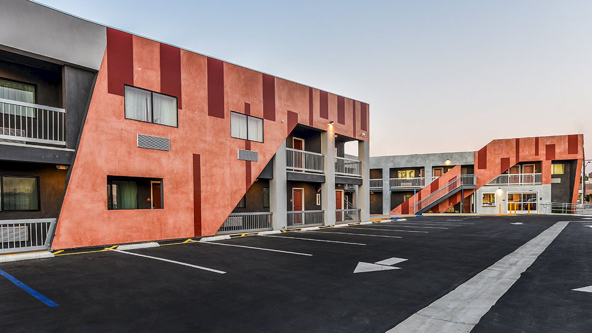 The image shows the exterior of a modern, two-story apartment complex with red geometric designs and an adjacent parking lot featuring marked spaces.