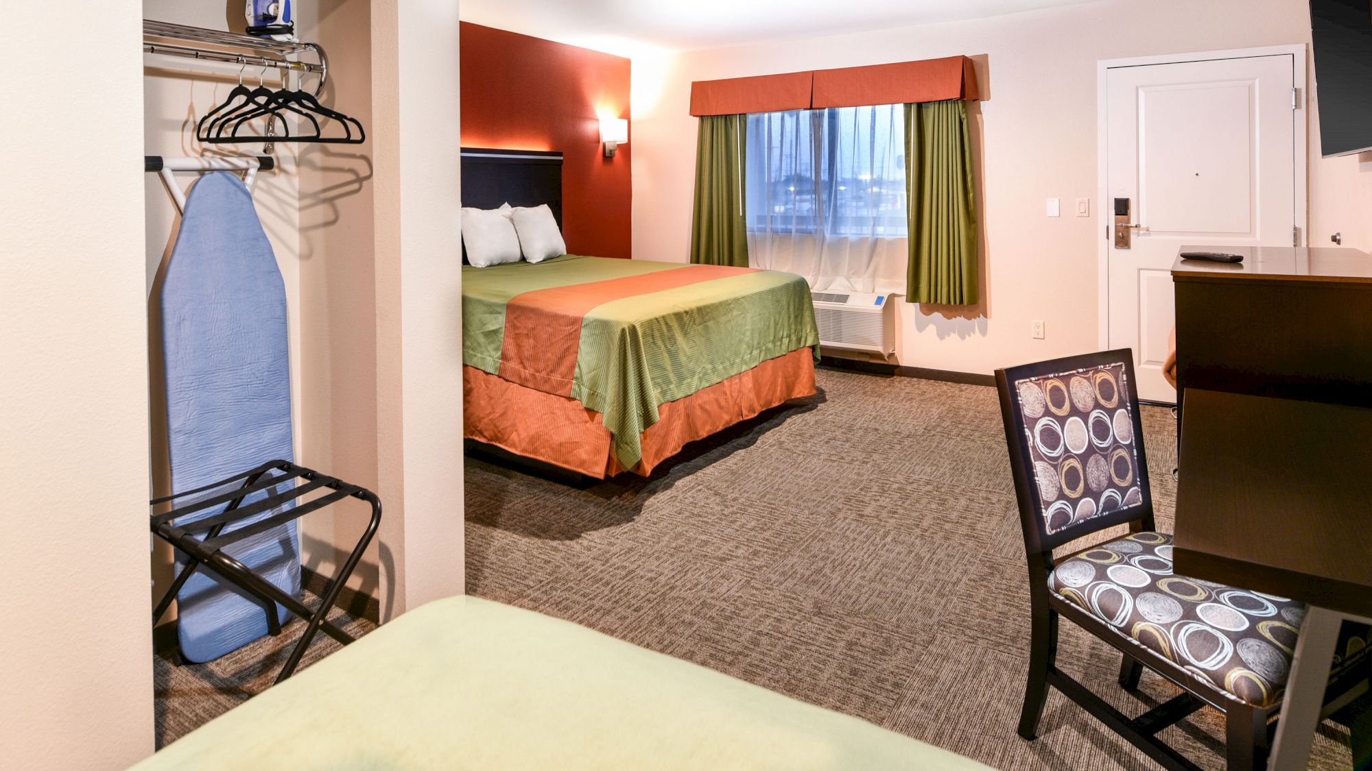 The image depicts a hotel room with a bed, an iron, an ironing board, a desk, a chair, and a luggage rack.