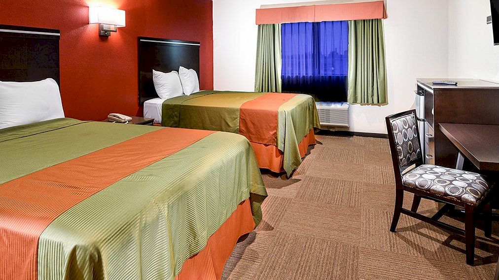 The image shows a hotel room with two beds, a work desk with a chair, a wall-mounted TV, and a window with green curtains.