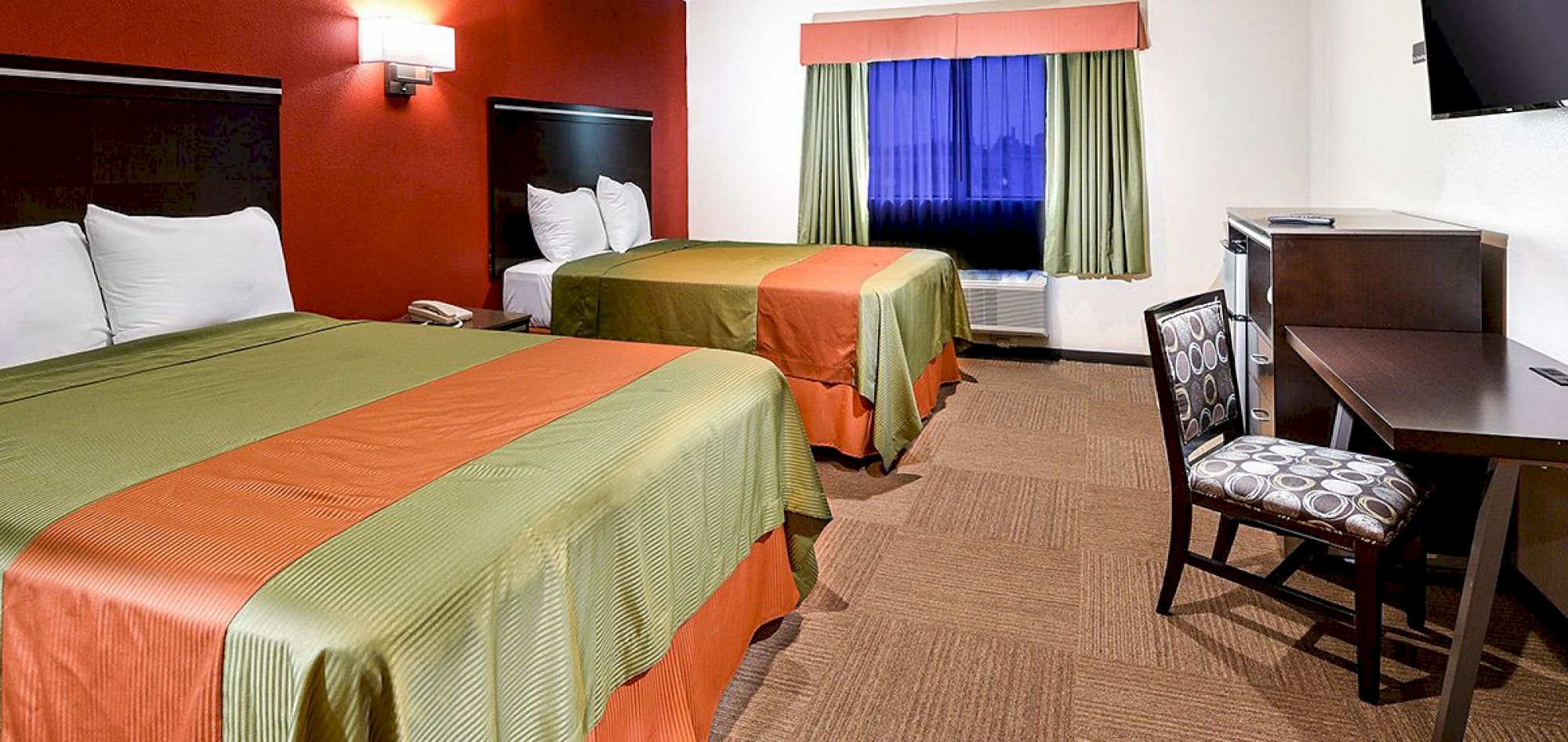 The image shows a hotel room with two beds, a work desk with a chair, a wall-mounted TV, and a window with green curtains.